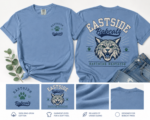 A blue short-sleeve graphic T-shirt shown from both front and back, laid flat with a knot on the left side and denim shorts partly visible on the right. The front has a small chest graphic reading Eastside Bobcats with a paw print; the back has a large bobcat head graphic with 'EASTSIDE Bobcats' and 'EASTSIDE BESTSIDE.