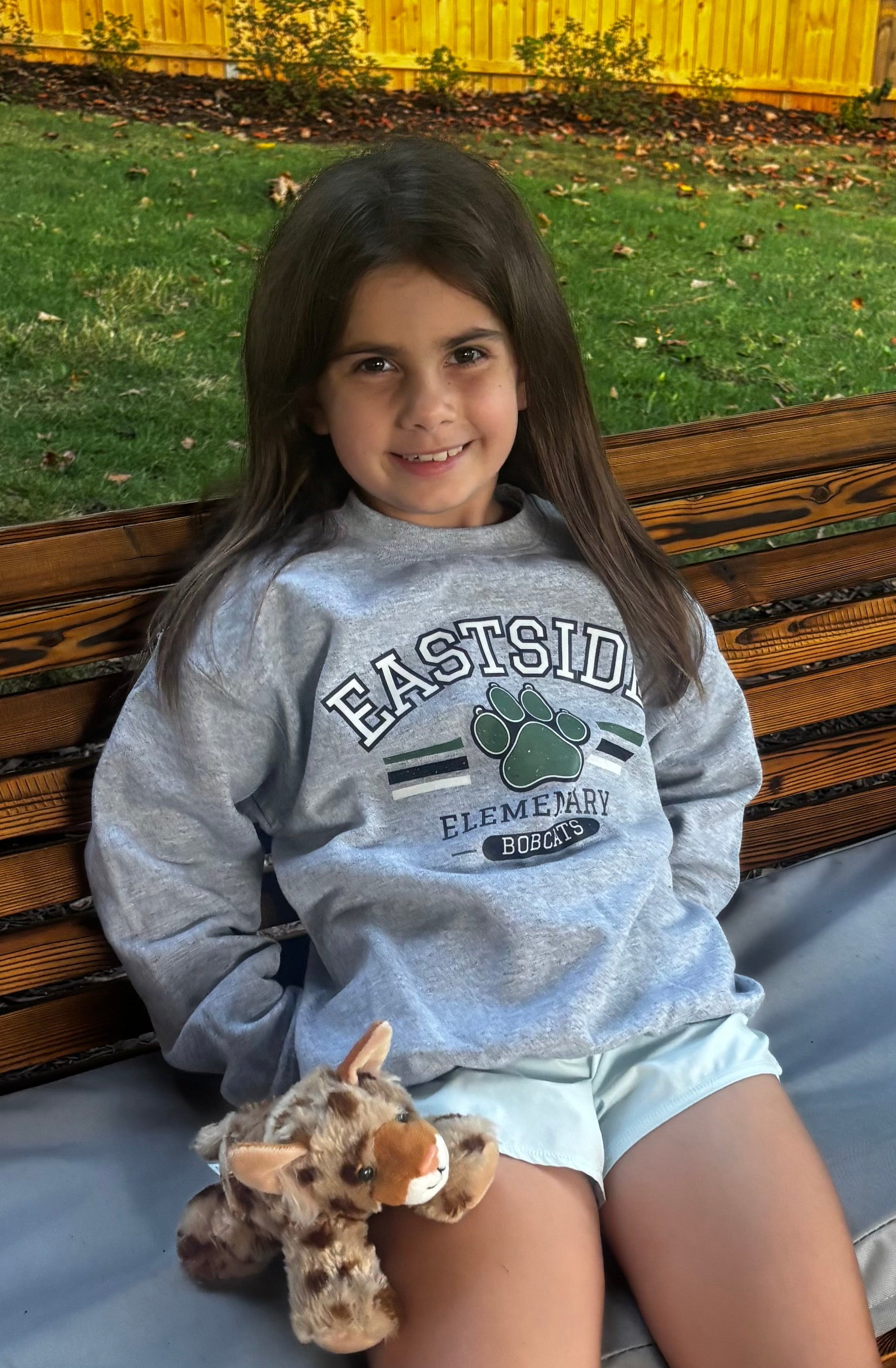 A young girl with long brown hair sits on a wooden park bench wearing a light gray sweatshirt with 'EASTSIDE ELEMENTARY BOYS' and a paw print graphic, paired with light blue shorts. She is holding a small stuffed Bobcat toy.